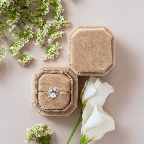 Beige jewelry box with a ring inside, surrounded by white flowers on a light background