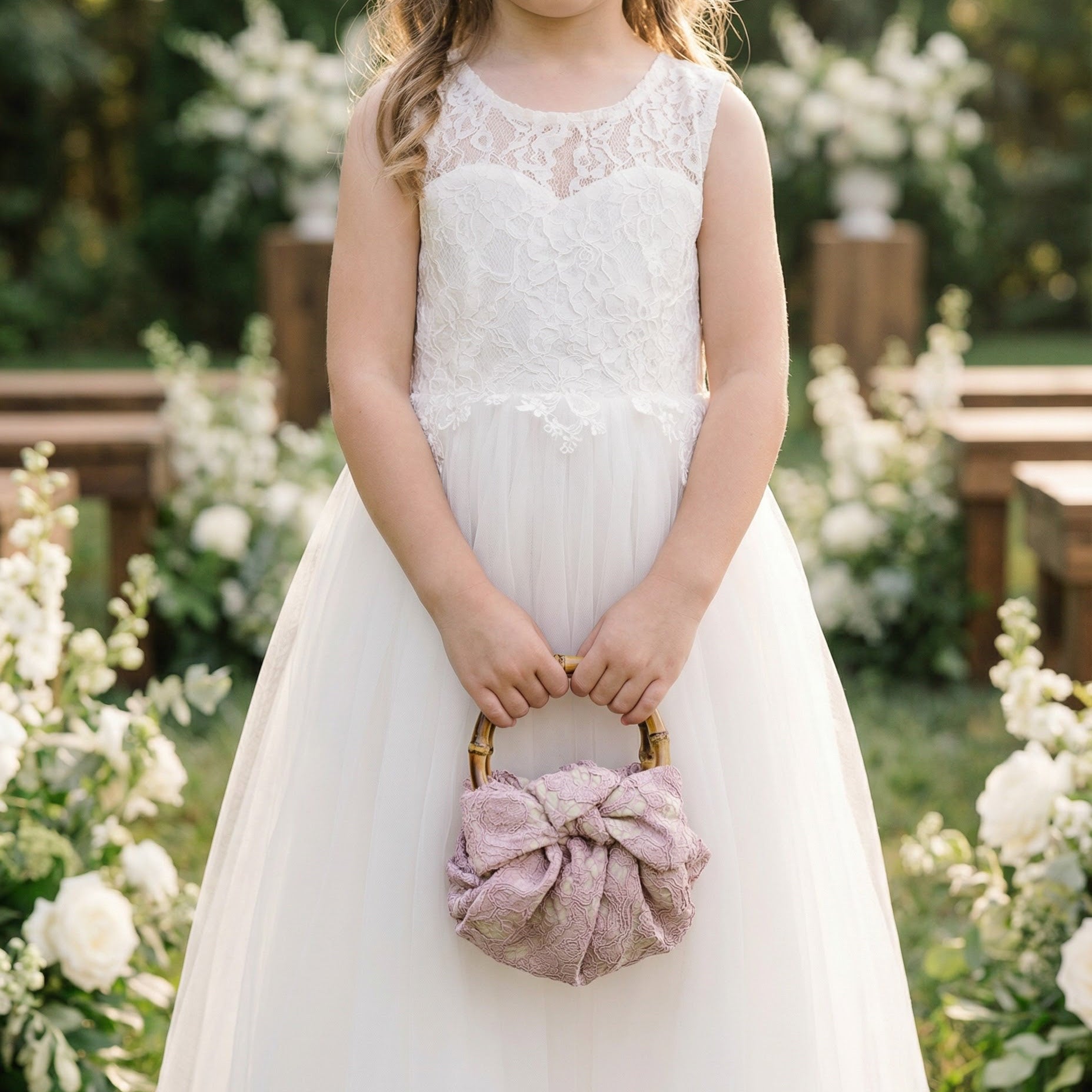Young girl in a white lace dress holding a pink flower basket in a floral setting.