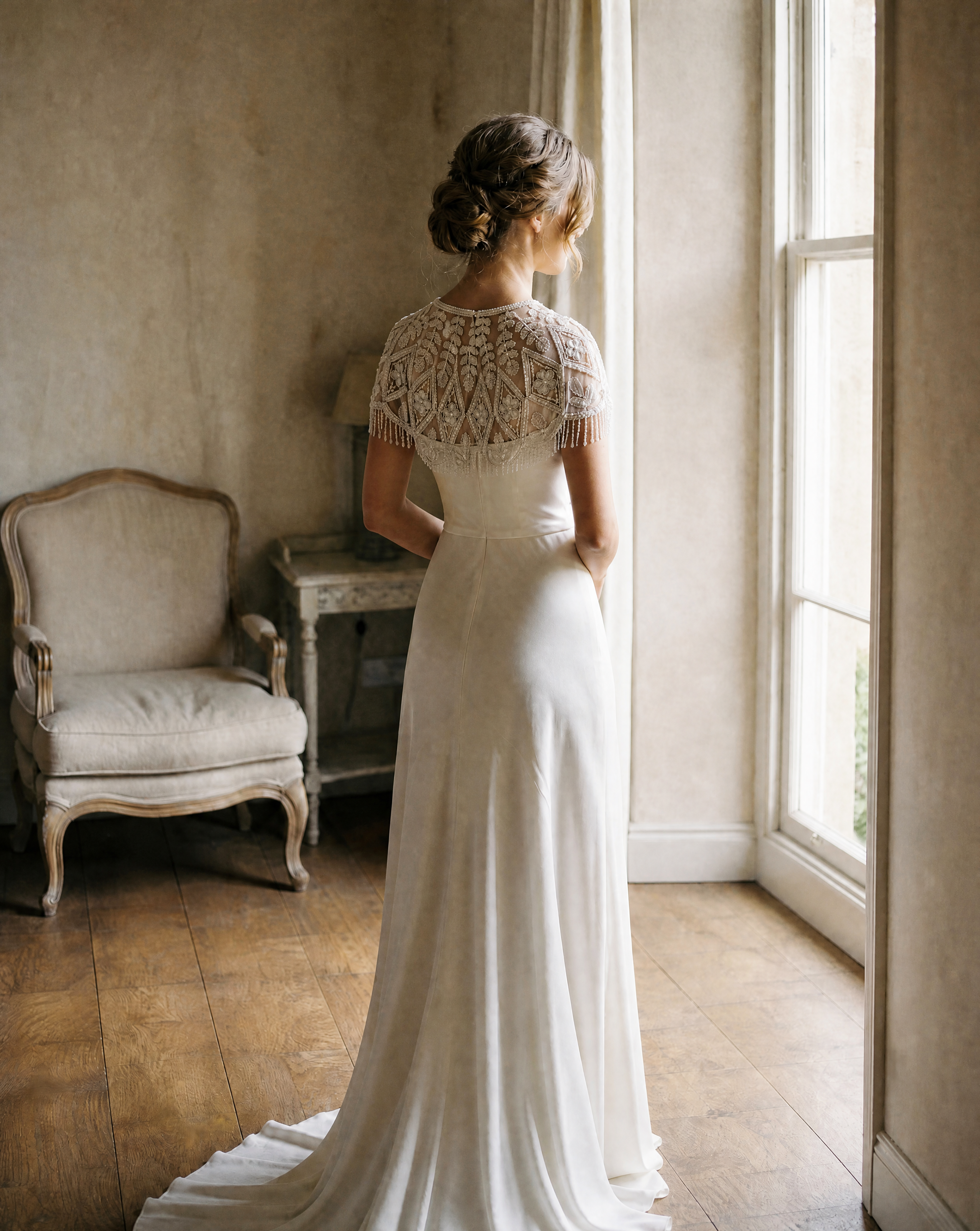 Woman in a white lace dress standing in a softly lit room with wooden floor and vintage chair.