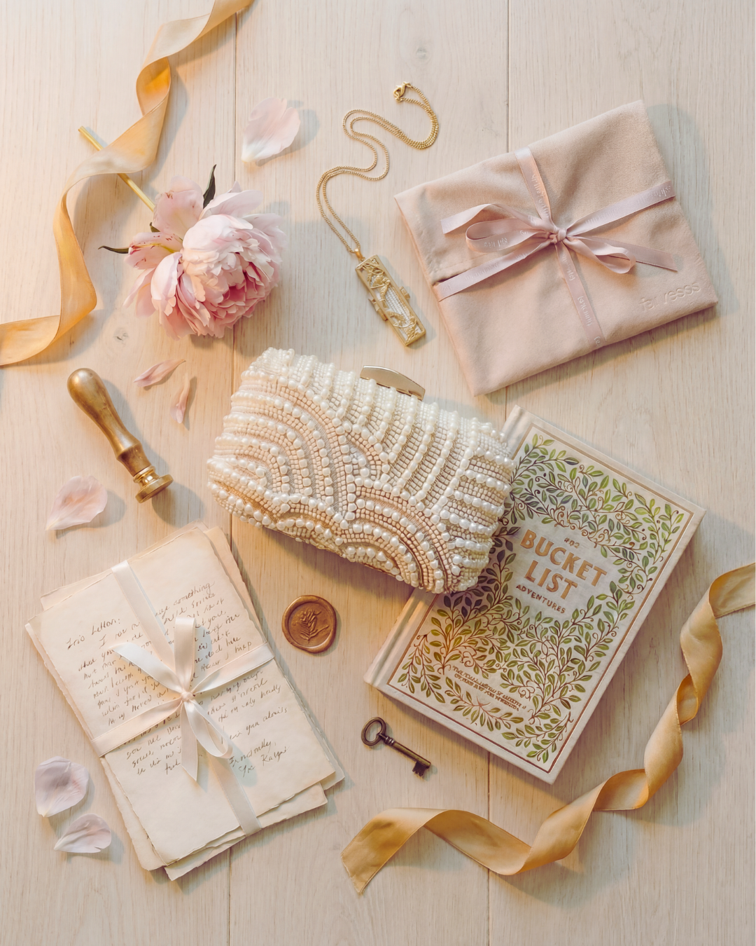 The Romantic wedding heirloom starter kit by For Keeps featuring an ivory beaded bridal clutch, Our Bucket List Adventures journal, gold locket, mauve suede pouch with For Keeps ribbon, handwritten love letters, brass wax seal, and blush peony on light French farmhouse wood