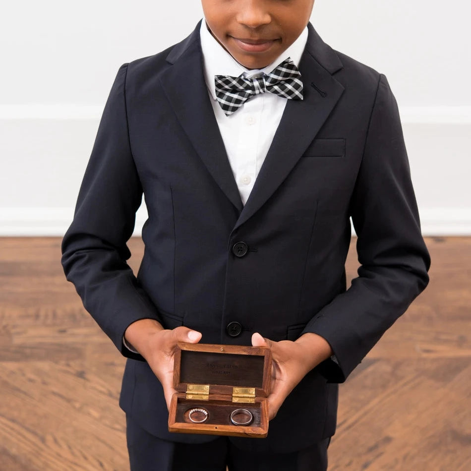 Young boy in a formal suit holding a wooden box with rings on a wooden floor.