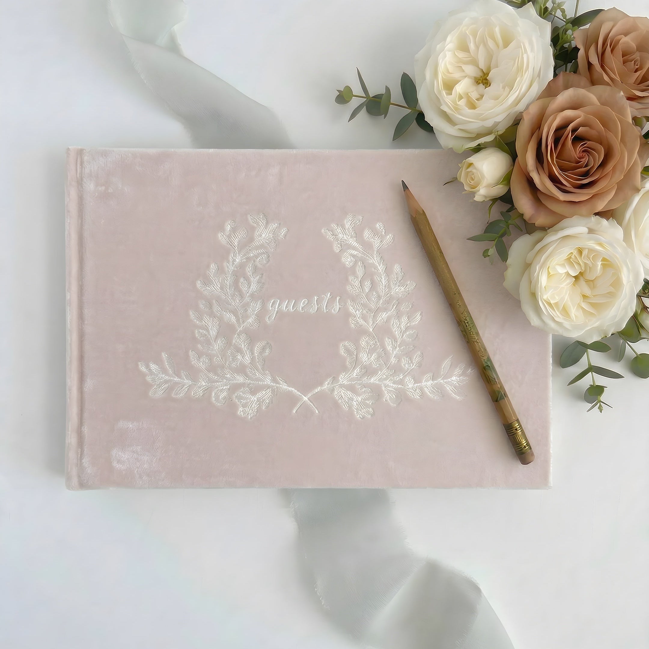 Pink guest book with floral design, pencil, and flowers on a white background