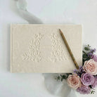 Beige embossed card with floral design on a white surface with pink and purple roses.