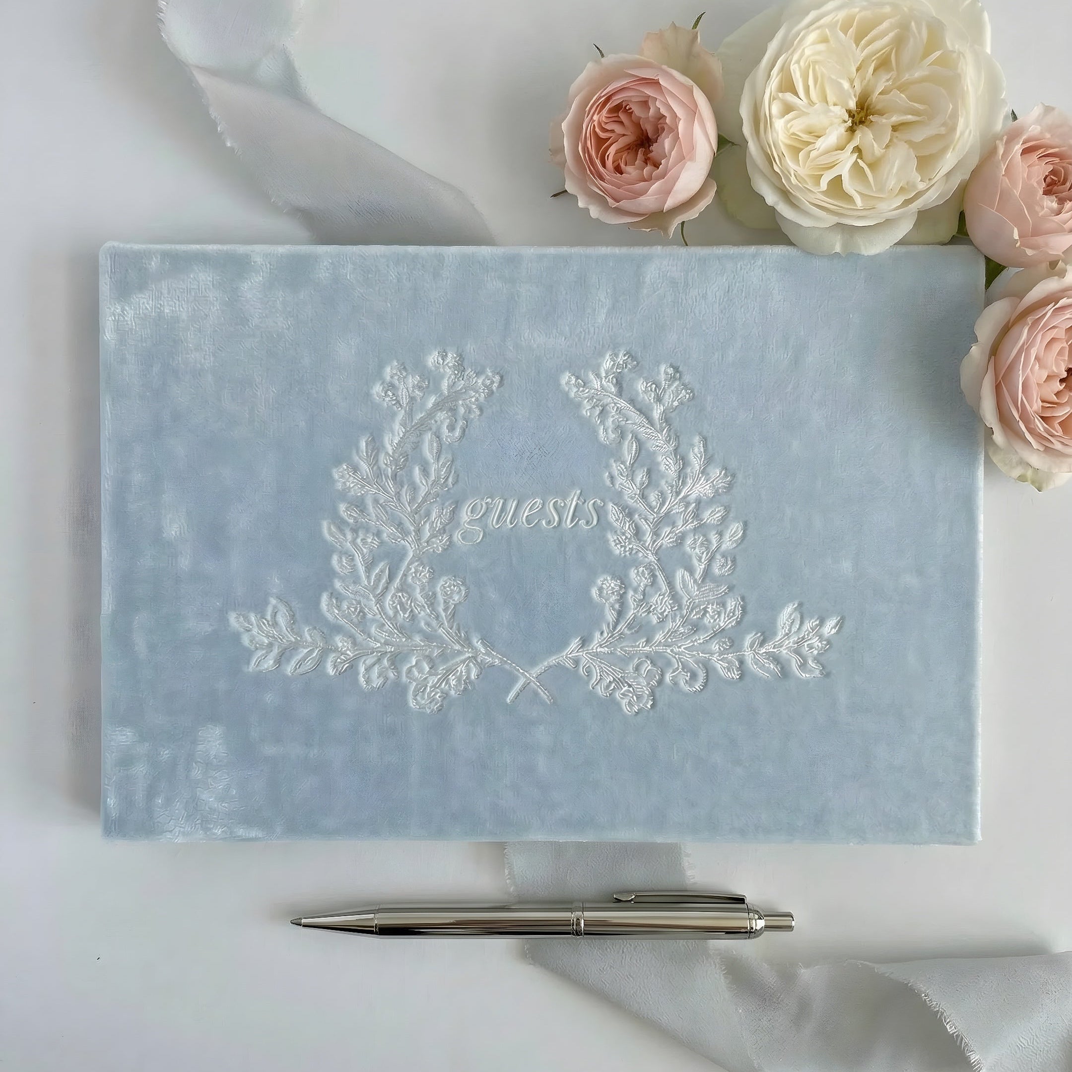 Light blue guest book with floral design and pen on a white surface with flowers.