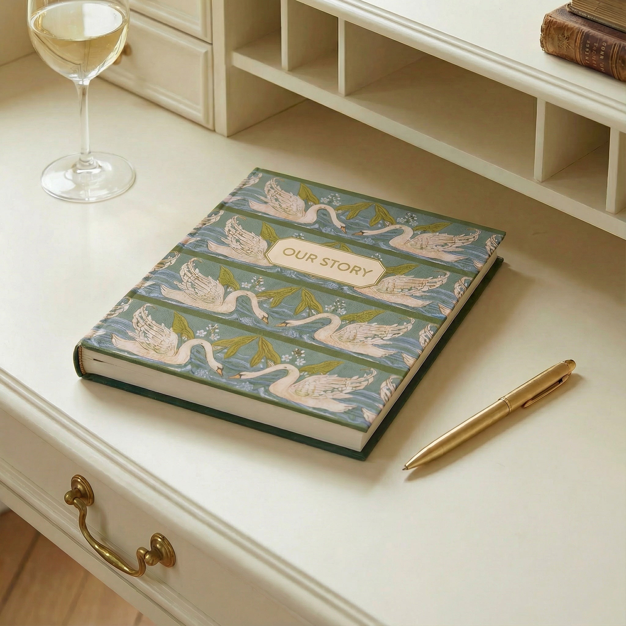 Decorative notebook with swan design and 'Our Story' text on a desk with a glass of wine and pen.