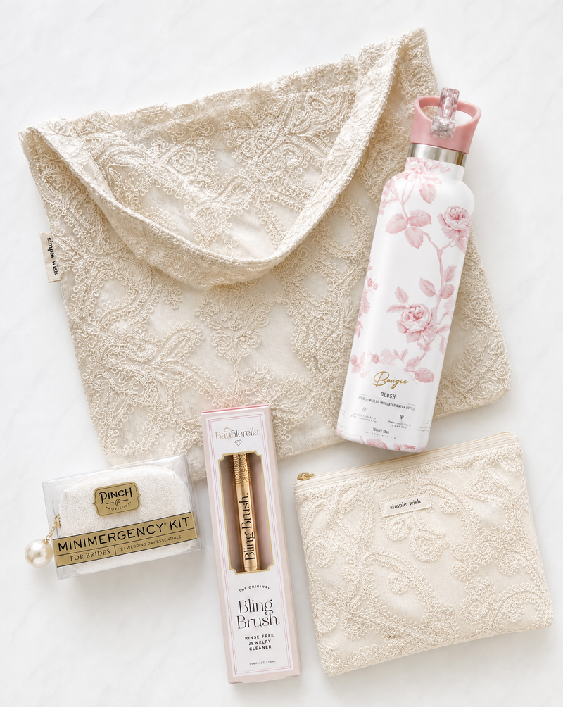 Collection of beauty products including a Boujee Bottle water bottle, bling ring cleaning brush, and bridal emergency kit on a heirloom lace tote.