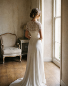 Woman in a white lace dress standing in a softly lit room with wooden floor and vintage chair.