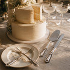 Two-tiered white wedding cake with decorative flowers on a table set with plates, cutlery, and glasses.