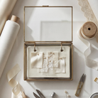 The Fabric Swatch Keep by For Keeps — a handmade brass and glass hinged keepsake box containing a linen-bound fabric swatch booklet with brass pin fasteners, shown open with wedding fabric swatches pinned inside, on dark ivory linen