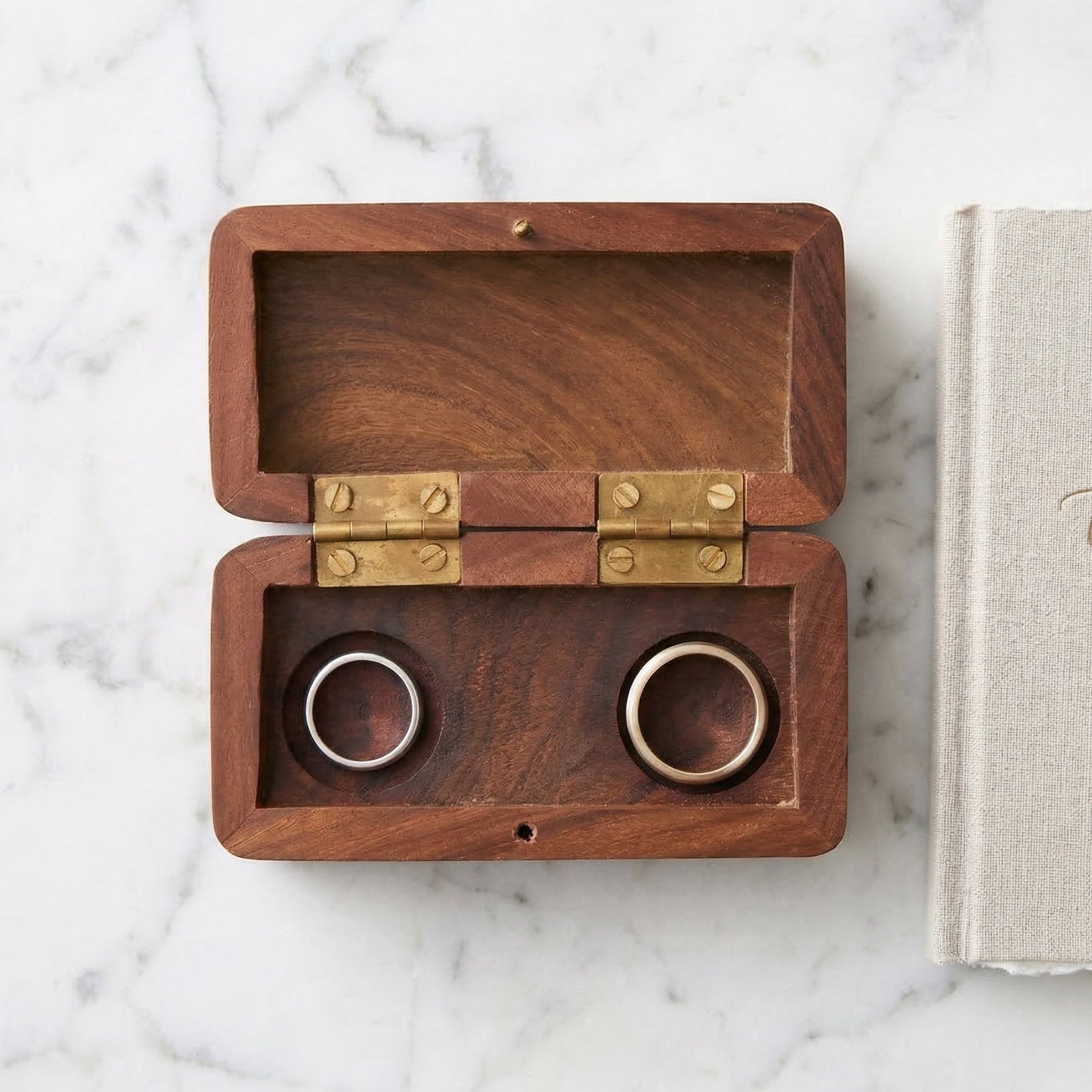 Wooden ring box with two rings inside on a marble surface next to a book titled 'Our Vows'.