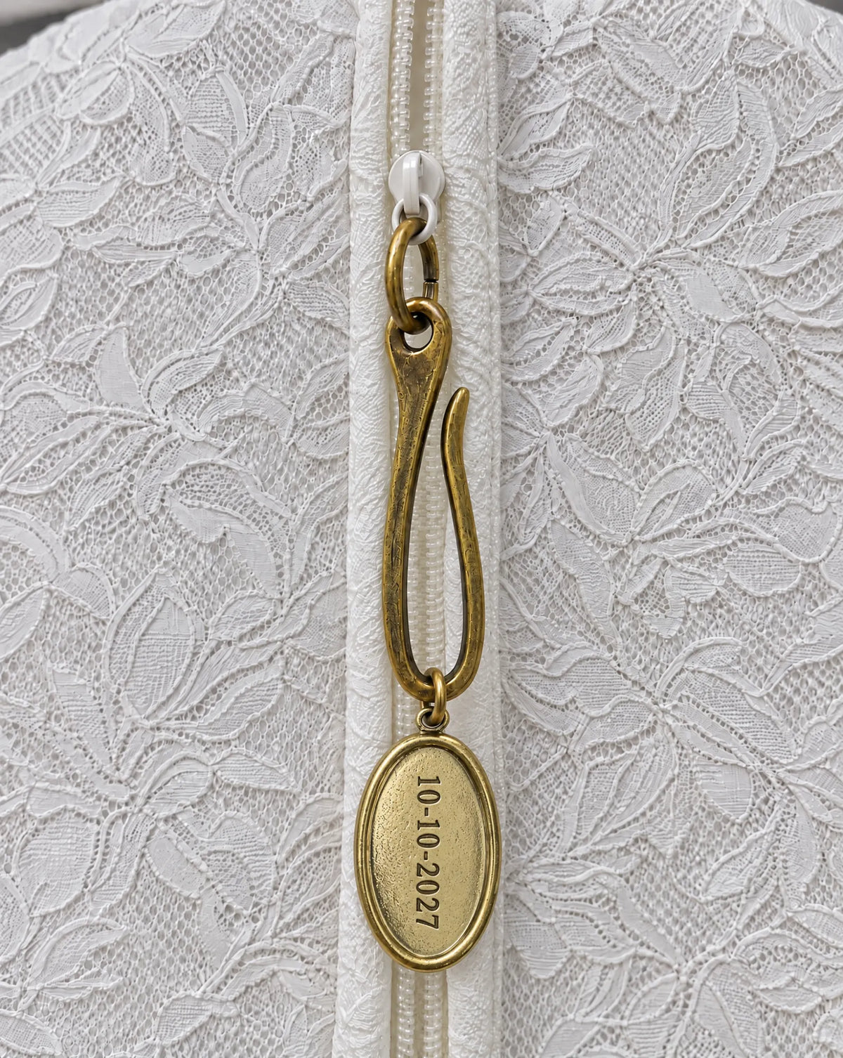 Brass wedding date charm on garment bag zipper: an antique brass hook and oval medallion hand-stamped 10-10-2027 on the ivory lace bridal garment bag