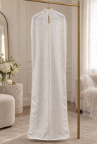 The Mariée ivory lace bridal garment bag hanging in a bridal suite, with a brass wedding date charm on the garment bag zipper